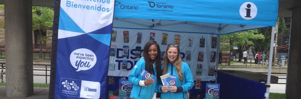 Two smiling tourist information specialists hold brochures in front of a mobile tourist information booth