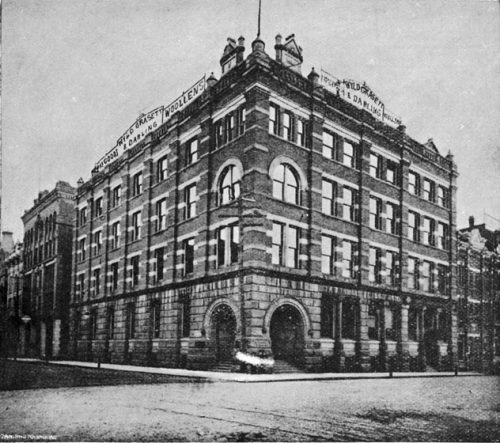 This four-storey building is built of cut stone on the first storey, and brick with stone details above. At street level, two stone arches, one on each wall at the corner of the building, lead to stairs to an outdoor vestibule, and then to the front door. Signs on top of each wall reads: Wyld Grasett and Darling, dry goods, woollens.