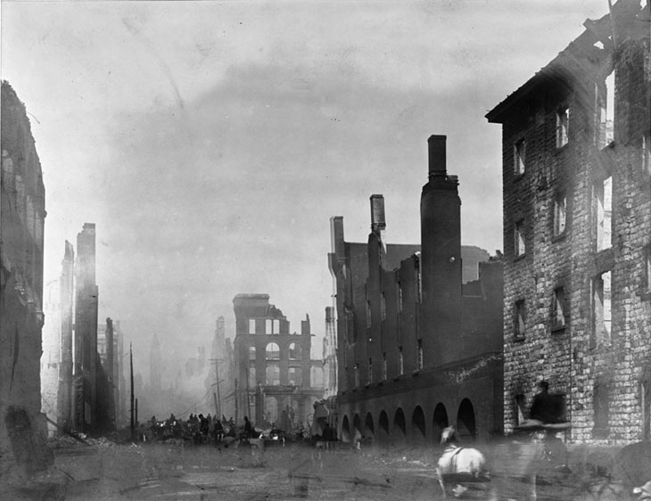 The stone shell of the mill and of other brick buildings line the foot of Bay Street. A white horse is pulling a carriage past the mill. Many people and horses are gathered farther up the street.