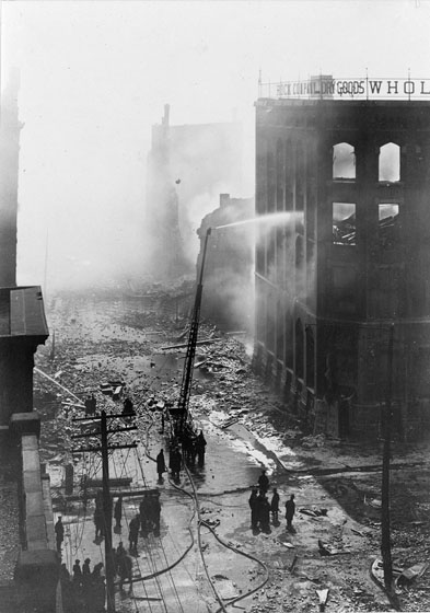 Firefighters use a water tower to spray the ruins of the Brock Building with water. The Bay Street façade of the building is intact, but smoke and rubble can be seen through the upper window openings.