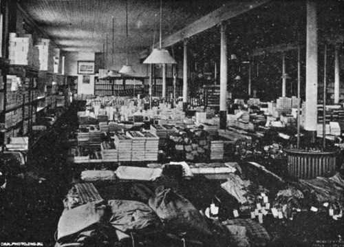 The long, wide room is punctuated by support pillars. Shelves along the walls and tables in rows filling the room are heaped with boxes and stacks of clothing and other goods.