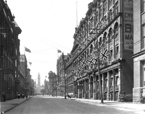 Stone and brick buildings on both sides of the paved street are three to five stories high. Many have arched windows and doorways, cut stonework, and other ornaments. Some modest lettered signs are hanging out over the concrete sidewalks from the buildings, but many stores have their names in raised lettering across the front of their buildings, or even painted on their sides. The clock tower of Old City Hall is visible in the distance at the end of the street.