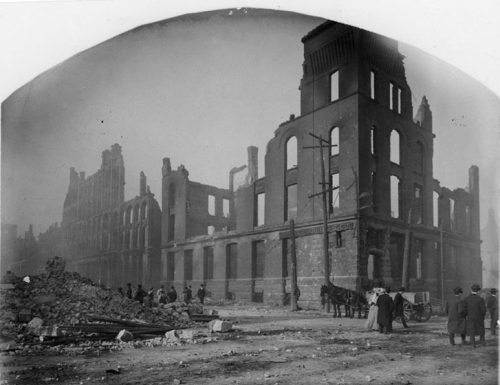 Most of the the ruins are several stories high. Pedestrians and a horse-drawn cart travel the streets, which are mostly clear. To the left, a building has completely collapsed, and cut stone blocks dot the brick rubble.