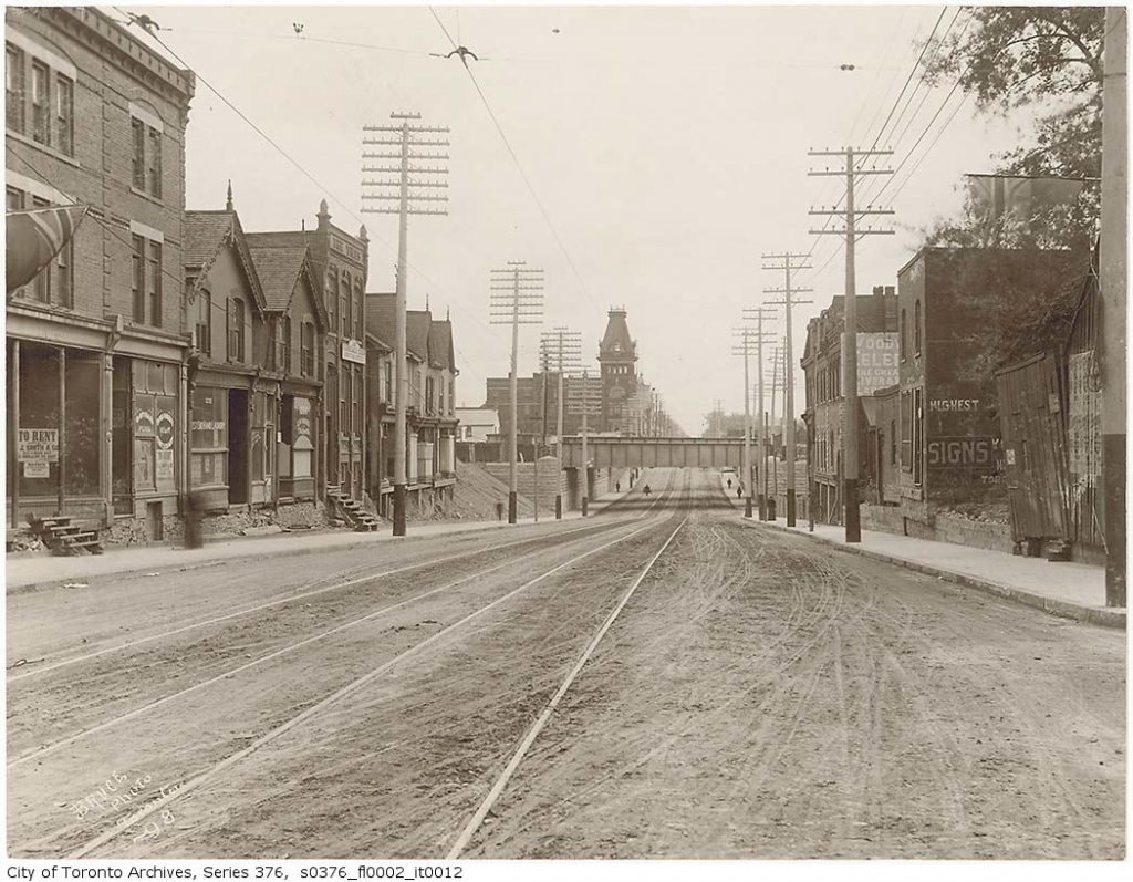 Queen Street is a dirt road with streetcar tracks, lined with stores, looking to bridge going over road