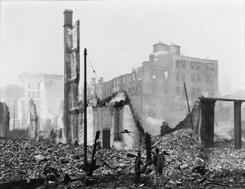 Two water tanks sit on top of a five-storey building that is damaged but still standing. The foreground is covered in rubble and the stubs of walls.