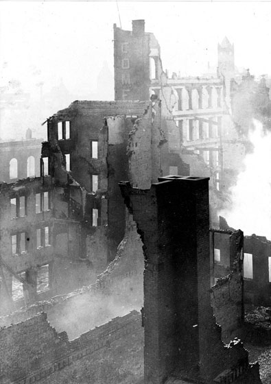 Ruined brick walls, a variety of heights, are viewed from above. Smoke is coming from ground level.