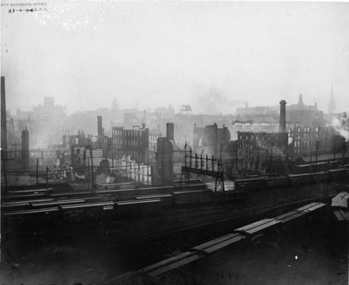 In the foreground are several railway tracks and trains. In the distance are the fire ruins, still smoking.