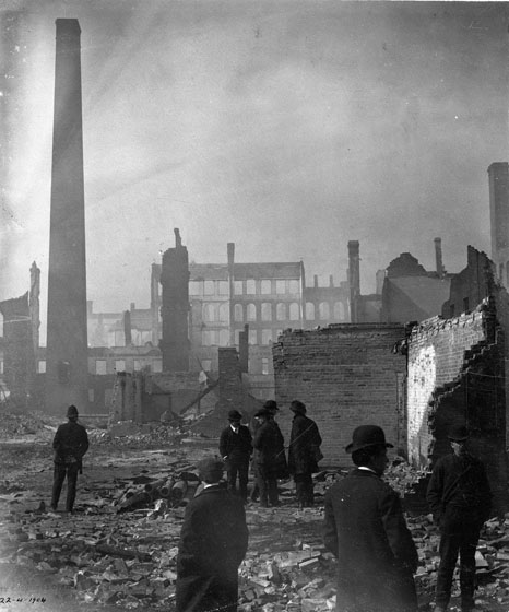 Men in business suits stand around in fire ruins, surrounded by partial brick walls. A tall chimney juts up at the left. In the distance stands a line of building facades with empty windows.