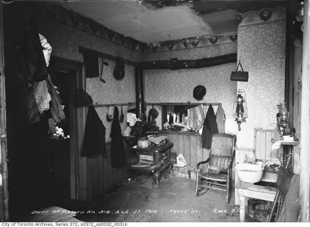 A crowded, dark room with wood-burning stove, table, chair, and lots of clothing hanging from the walls.