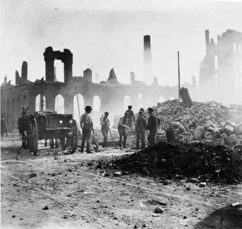 Eight men are preparing to clear a fire ruin site by loading bricks and cut stone into a horse-drawn cart. A few men have shovels. Another seems to be picking up a piece of stone with his hands.