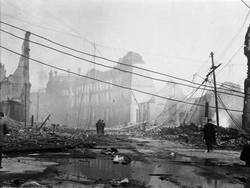 Ruins and facades with nothing behind them line the street. Wires tangle above street level and lie in strands on the ground. Bricks and burnt timbers litter the street. A wide puddle of water lies on the street in the foreground.