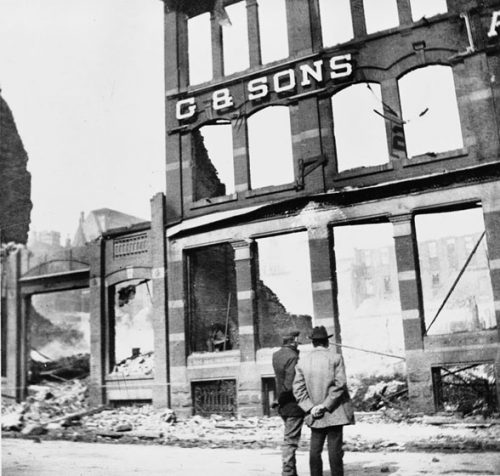 Two men stand on the street, looking at the ruins of a building. At least three stories of the brick façade still stand, with empty door- and window-frames. On front of the building are the letters G & SONS.