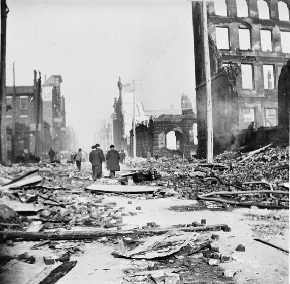 Men walk down the street, away from the camera, surrounded by fire ruins. The street is choked with bricks, timbers, discarded fire hoses, and other litter.