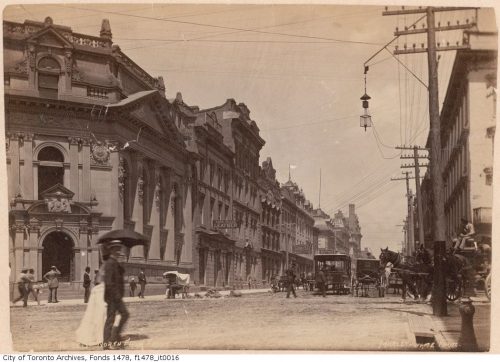 People crossing road, with grand three-storey stone and brick buildings lining the street.