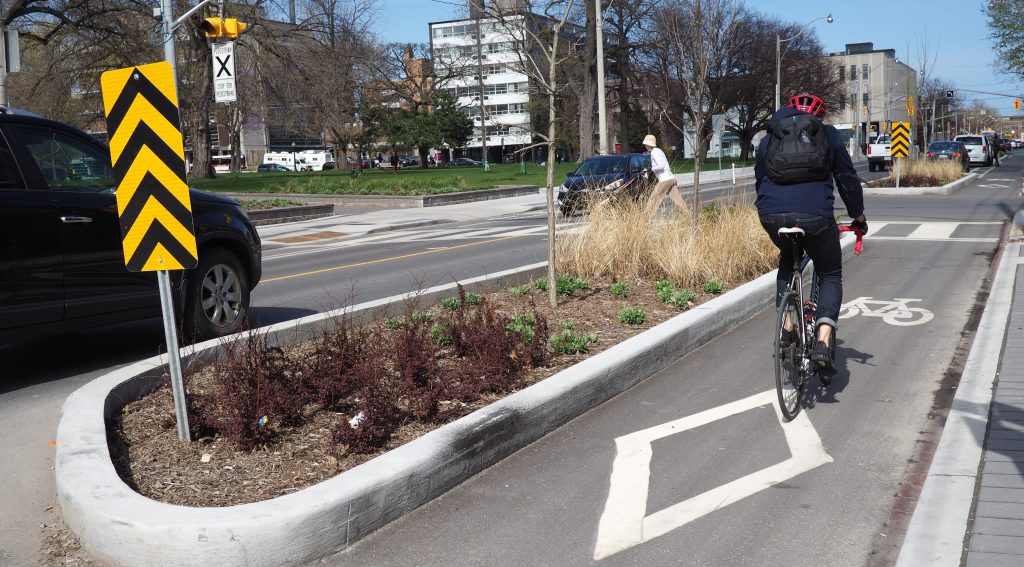 Cyclist using the cycle track on Gerrard St E separated by a permanent planter. Background has a pedestrian crossing with a pedestrian pushing a stroller across.