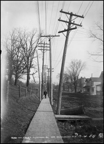 A man walks down a plank sidewalk beside a muddy road. Hydro poles with many wires line both sides of the sidewalk.