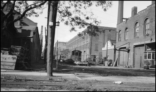 Two cylindrical lanterns on a lamp post surrounded by large brick industrial buildings.