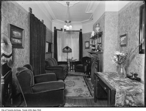 Narrow living room with plush chairs, fireplace, electric overhead light and floor lamp.