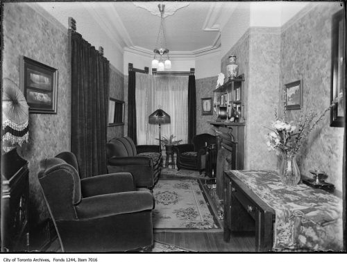 Narrow living room with plush chairs, fireplace, electric overhead light and floor lamp.