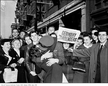 A crowd watches an Air Force pilot and a woman kissing while another woman holds up a newspaper with the headline Germany Surrenders.