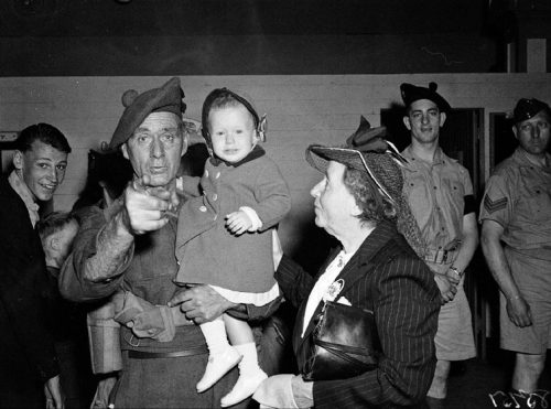 An older man in military uniform holding a little girl points at the camera.