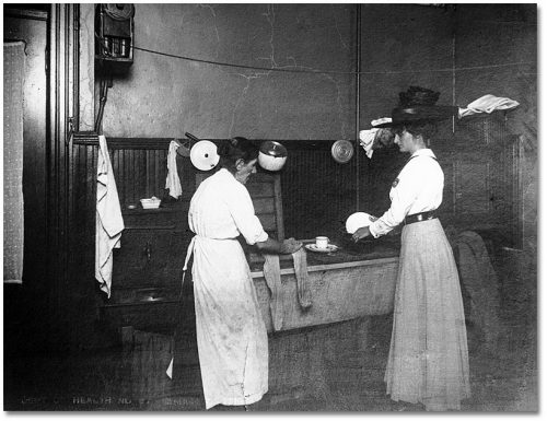 Two women wash dishes in a large rectangular sink.