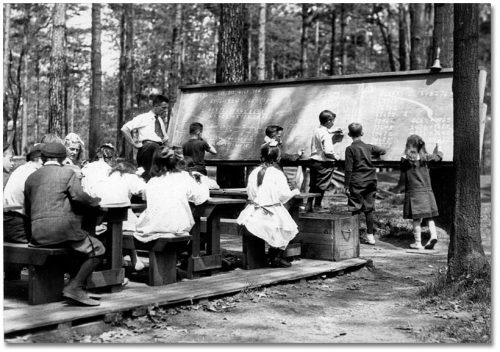 In a clearing in the trees, children sit at desks in front of a blackboard.