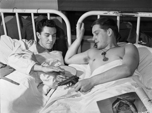 Two young men lie in hospital beds. One is displaying a manicure set, the other a pistol.