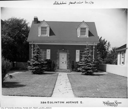 An ordinary-looking storey and a half house with a manicured lawn and evergreens in front.