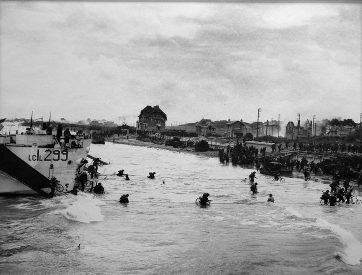 Soldiers carrying bicycles disembark from a troop carrier and wade through the water to a beach. Bombed-out houses line the beach.