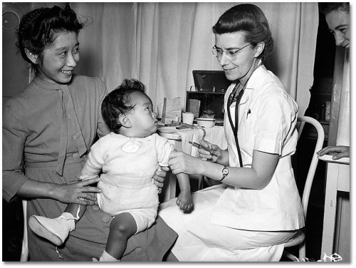 A woman of Asian heritage holds her toddler on her lap while a nurse or doctor prepares to give the child a needle.