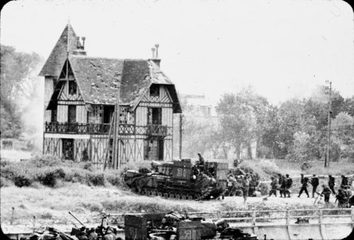 A tank leads a line of soldiers past a Tudor-style house. Its roof is damaged, possibly by the fighting. More houses and trees are in the backrground. More soldiers are in the foreground, just coming into the picutre.