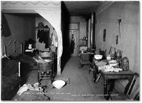 A dark, cluttered room with a wood-burning stove and a table against one wall and beds against the other.