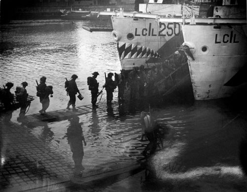 Soldiers, seen in silhouette, walk across narrow wooden gangplanks to troop carriers (ships), one of which has a mouth with sharp teeth painted on its bow.