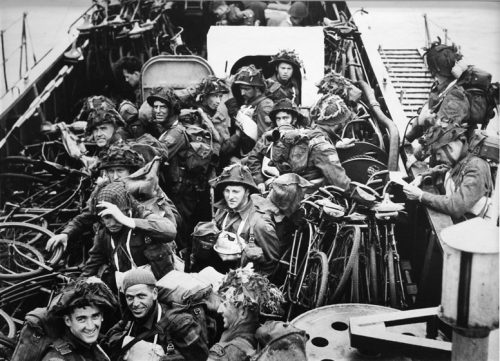 Soldiers and bicycles are packed close together on the narrow deck of a troop carrier. They are wearing helmets and backpacks, and some have equipment hanging across their chests as well.