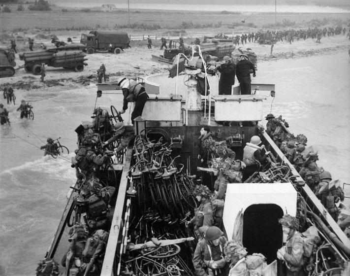 Soldiers carrying bicycles disembark from a troop carrier and wade through the water to a beach, where they are lining up. A truck, a tank, and trailers loaded with ammunition stand on the beach. The land beyond the beach is flat.