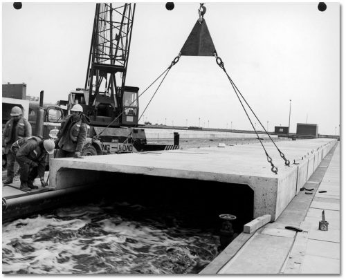 A crane lowers a large C-shaped concrete cover over a basin of roiling water.