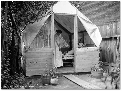 Nurse stands beside a man in a bed in a tent.