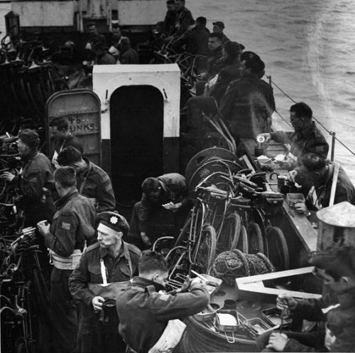 Soldiers are eating a meal while standing on the narrow deck of a troop carrier, surrounded by tightly packed bicycles. They are balancing their dishes on bicycles, railings, spools of rope, and other items on deck. They are eating out of rectangular metal containers.