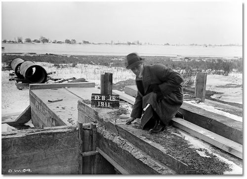 Man in winter coat squats on low block concrete structure and holds ruler against a large crack.