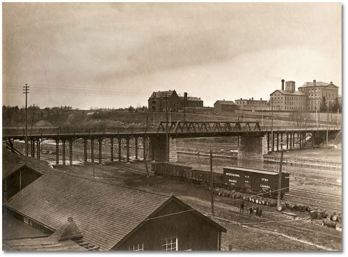 Bridge and railway tracks in the foreground, with a three-storey brick building in the back centre, and the stone Don Jail to the right.