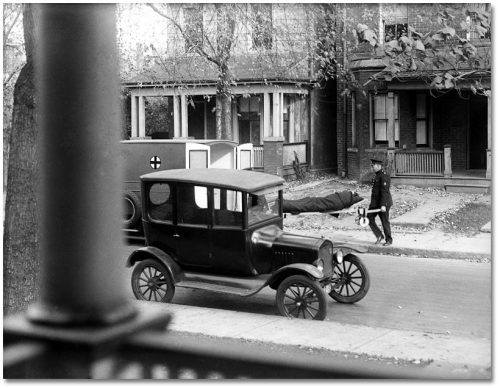 On a residential street, two men carry a blanket-covered person on a stretcher into the open back doors of a motorized ambulance.