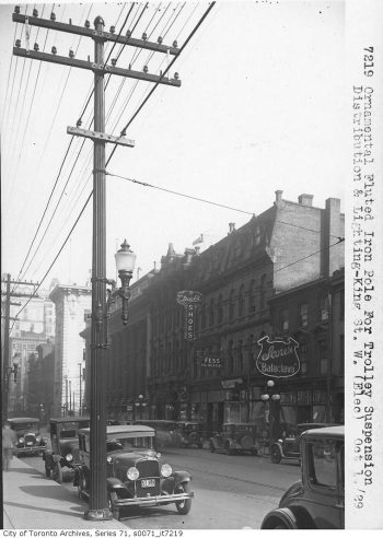 Street lined with stores and parked cars. In the foreground is a hydro pole with a street lamp on the side.