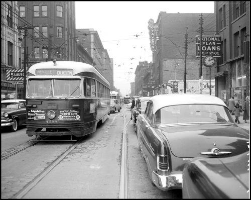 A streetcar with a rounded roof on one side of the street and large 1950s cars on the other.