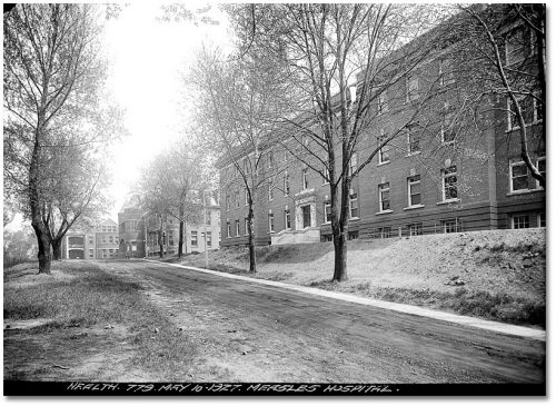 Dirt road lined with trees, with a three-storey blocky brick building on one side, and an older brick building with turrets and verandahs at the end of the road.