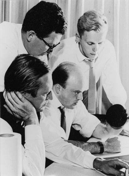 Group of white male architects in white shirts and ties gathered around desk