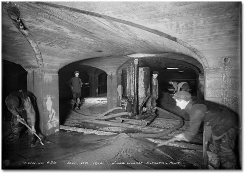 Men spray water from hoses onto piles of sand in a low-ceilinged vaulted reservoir.