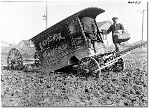 Men in muddy boots unloading baskets of bread from a mud-covered delivery wagon that has come off its wheels.