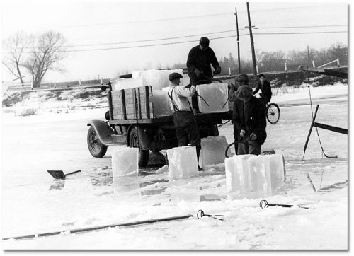 On a frozen lake, men use metal tongs to lift two-foot-wide cubes of ice onto the back of a truck.