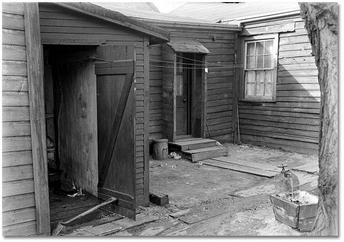 Courtyard surrounded by small wooden houses, with water running from an outdoor tap into a square wooden box.